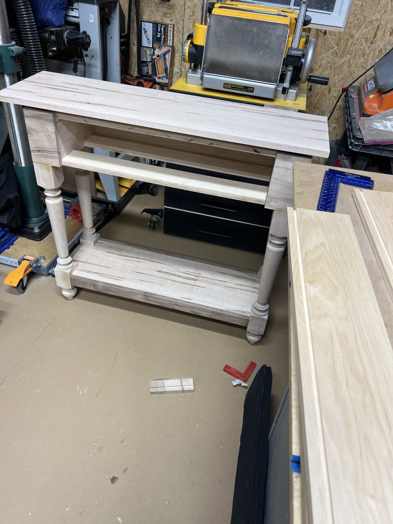 The table coming together in the shop before finish — legs, case and lower shelf assembled, drawer opening visible at the front.