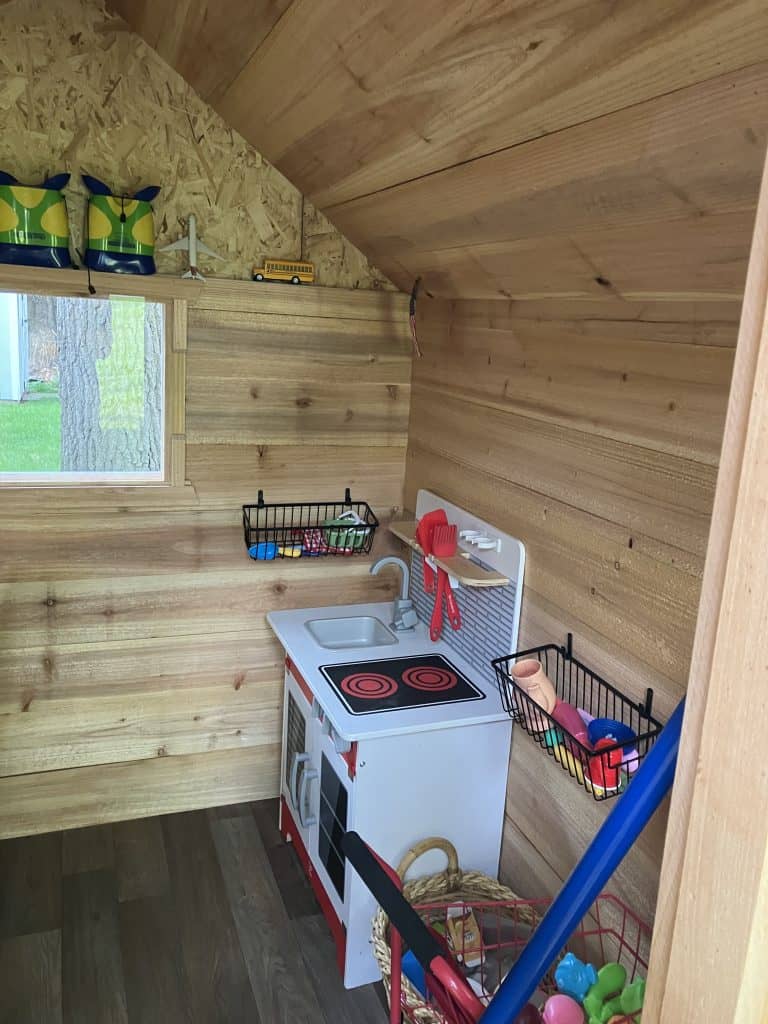 The other corner of the interior, with a toy kitchen, wire baskets hung on the cedar walls and a shelf tucked under the roof slope.