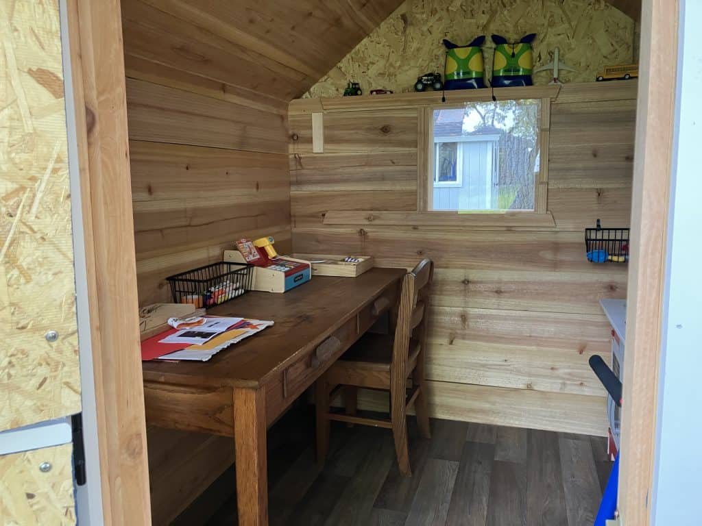 Inside, the cedar picket board walls and vinyl plank floor, with a small desk and chair under one of the plexiglass windows.