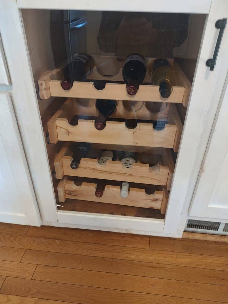 The four pine racks pushed back into the cabinet opening that was originally built for a wine cooler, each rack notched to cradle the bottles.