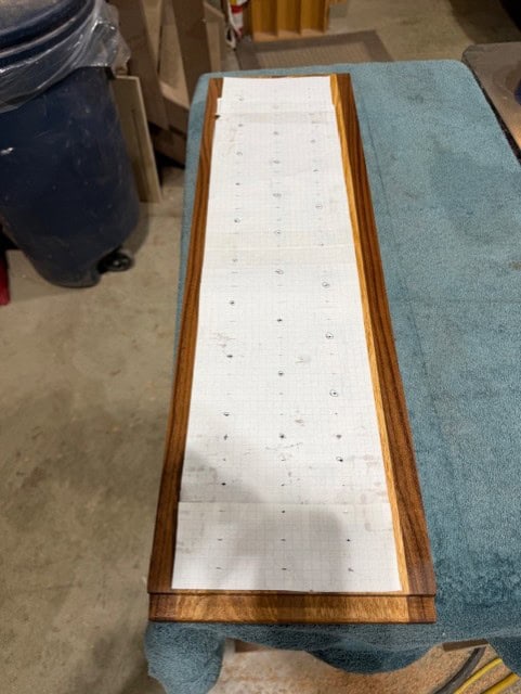 A gridded paper template taped to a walnut-edged board, used to lay out the hole pattern.